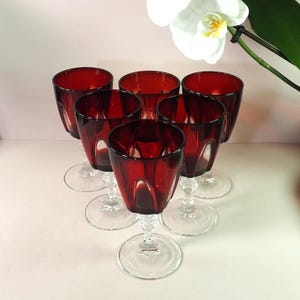 Può includere: Sei bicchieri rosso rubino con steli e basi trasparenti e ornati. I bicchieri sono disposti a cerchio. Fiori di orchidea bianchi sono visibili sullo sfondo. I bicchieri sono probabilmente per vino o acqua.