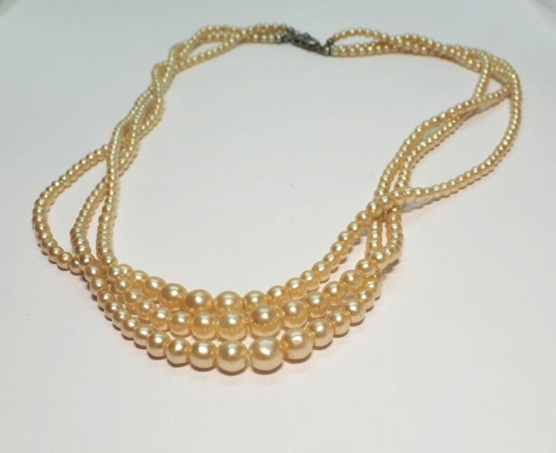 Puede incluir: Un collar vintage con tres hilos de perlas de imitaci&oacute;n de color crema. Los hilos est&aacute;n entrelazados en la parte superior y asegurados con un cierre plateado. El collar tiene un dise&ntilde;o cl&aacute;sico y elegante, perfecto para eventos.