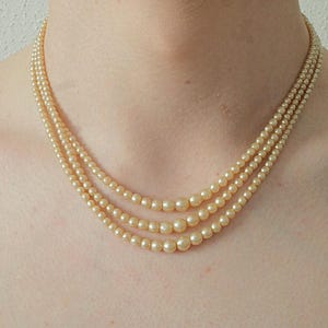 Puede incluir: Un collar de perlas de tres hilos con perlas de color crema de diferentes tama&ntilde;os. El collar est&aacute; colocado alrededor del cuello de una persona, mostrando los tama&ntilde;os graduados de las perlas. Las perlas tienen un brillo sutil.