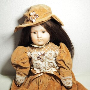Könnte beinhalten: Eine Vintage-Puppe mit langen braunen Haaren, die einen Cordhut und ein Kleid in Beige- und Brauntönen trägt. Das Kleid hat Spitze und eine dekorative Brosche. Die Puppe hat ein Porzellangesicht mit bemalten Gesichtszügen.