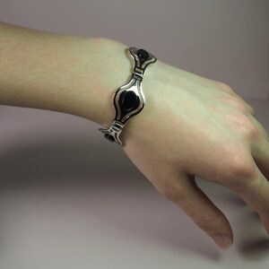 May include: A silver-toned bracelet with black oval-shaped accents. The bracelet is worn on a wrist, showcasing its design. The bracelet's design features a series of connected, stylized oval shapes with black centers, creating a modern look.
