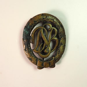 May include: A gold-toned metal pin with a wreath of leaves surrounding the letters "DB" in a stylized font. The pin has a bow at the bottom.