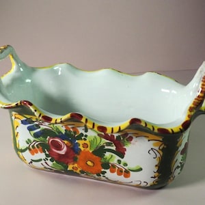 Puede incluir: Un plato de servir de cerámica blanca con dos asas. El plato tiene un borde festoneado y está decorado con diseños florales verdes, amarillos, rojos y azules.