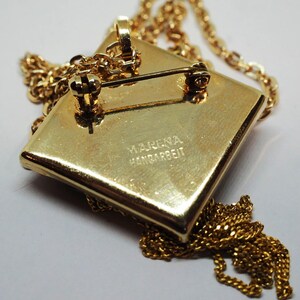 May include: A gold-toned pendant necklace with a square pendant. The pendant has a pin on the back, suggesting it can also be worn as a brooch. The chain and pendant are a matching gold color. The pendant has the text "MARENA HANDARBEIT".