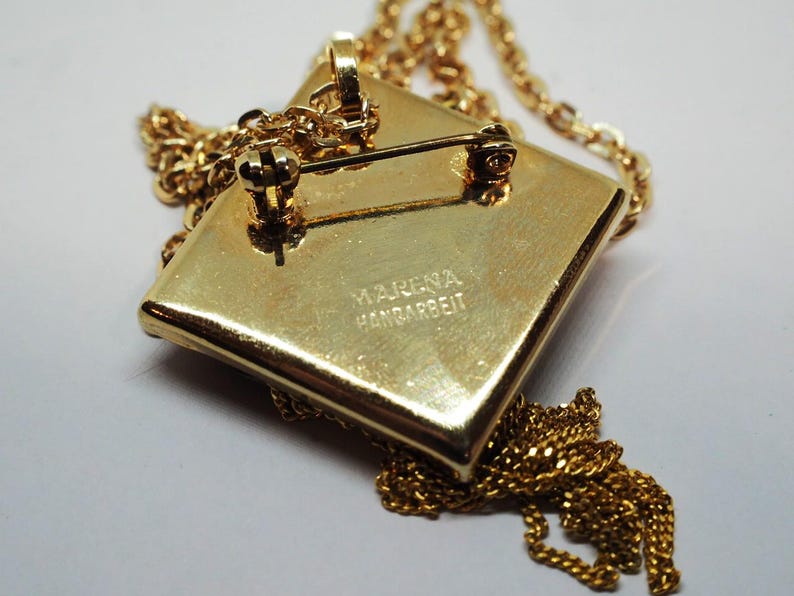 May include: A gold-toned pendant necklace with a square pendant. The pendant has a pin on the back, suggesting it can also be worn as a brooch. The chain and pendant are a matching gold color. The pendant has the text "MARENA HANDARBEIT".