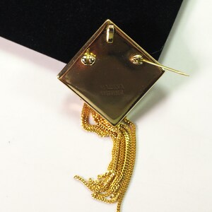 May include: A gold-toned, diamond-shaped brooch with a dangling chain tassel. The back of the brooch features a pin clasp and the inscription "H. VERNA SANDBERG". The tassel is made of multiple strands of delicate chain.