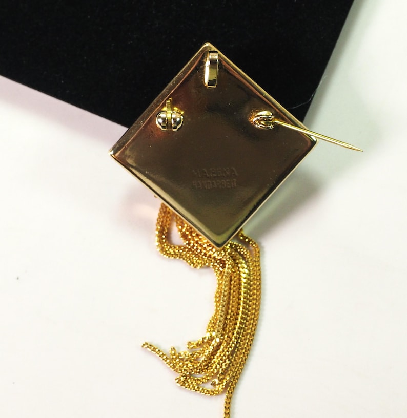 May include: A gold-toned, diamond-shaped brooch with a dangling chain tassel. The back of the brooch features a pin clasp and the inscription "H. VERNA SANDBERG". The tassel is made of multiple strands of delicate chain.