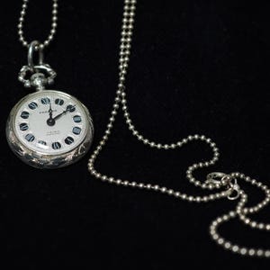 Può includere: Ciondolo orologio da tasca argentato con quadrante bianco, numeri romani neri e lancette nere. L'orologio è attaccato a una collana a catena a sfera argentata. La cassa dell'orologio ha un design ornato e inciso.