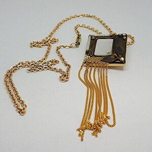 May include: Gold-toned necklace featuring a square pendant with a mirrored center and decorative leaf accents. The pendant is adorned with small crystals and dangling chains. The necklace has a delicate chain.
