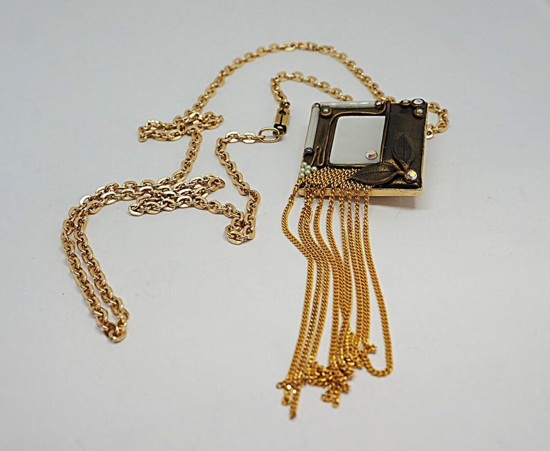 May include: Gold-toned necklace featuring a square pendant with a mirrored center and decorative leaf accents. The pendant is adorned with small crystals and dangling chains. The necklace has a delicate chain.