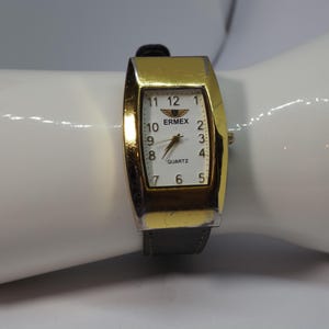 Puede incluir: Un reloj de pulsera dorado con esfera rectangular y correa de cuero negro. La esfera del reloj es blanca con números y manecillas negras, y se muestra la marca "ERMEX". El reloj mide aproximadamente 3,8 cm de largo.