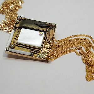 May include: A gold-toned necklace with a square pendant featuring a white, rectangular center. The pendant is adorned with small pearls, crystals, and leaf accents. A cascade of gold chains hangs from the pendant.