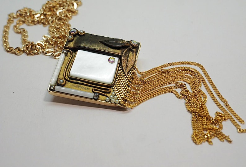 May include: A gold-toned necklace with a square pendant featuring a white, rectangular center. The pendant is adorned with small pearls, crystals, and leaf accents. A cascade of gold chains hangs from the pendant.