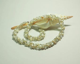 May include: A delicate necklace and matching bracelet featuring white pearls and clear crystals. The jewelry is draped over a large, cream-colored seashell, creating a coastal theme. The necklace is approximately 16 inches long.