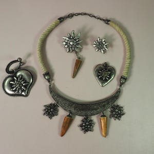 May include: A silver necklace with a rope cord and a crescent moon pendant with edelweiss flower accents. The necklace comes with a pair of edelweiss flower earrings and a heart-shaped pendant with an edelweiss flower.