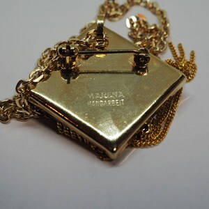 May include: A gold-toned pendant necklace with a square, textured pendant. The pendant has a pin on the back and is attached to a delicate chain. The text "MARENA HANDARBEIT" is stamped on the back of the pendant.
