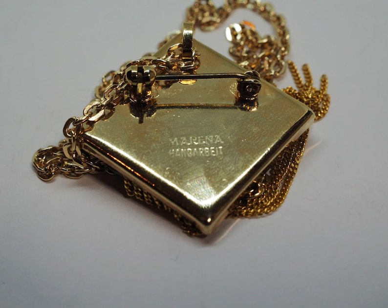 May include: A gold-toned pendant necklace with a square, textured pendant. The pendant has a pin on the back and is attached to a delicate chain. The text "MARENA HANDARBEIT" is stamped on the back of the pendant.