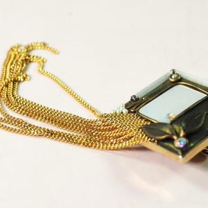 May include: A gold-toned brooch with a rectangular frame and a white center, adorned with small gemstones and a leaf design. Multiple strands of gold chain cascade from the brooch, creating a layered effect. The jewelry piece is set against a light pink background.