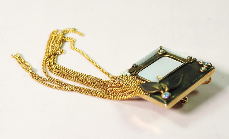 May include: A gold-toned brooch with a rectangular frame and a white center, adorned with small gemstones and a leaf design. Multiple strands of gold chain cascade from the brooch, creating a layered effect. The jewelry piece is set against a light pink background.