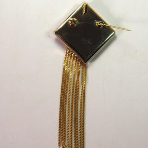 May include: A gold-toned brooch with a diamond-shaped top and dangling chains. The brooch has a pin closure on the back. The chains are made of small, linked segments. The brooch is likely vintage or costume jewelry.