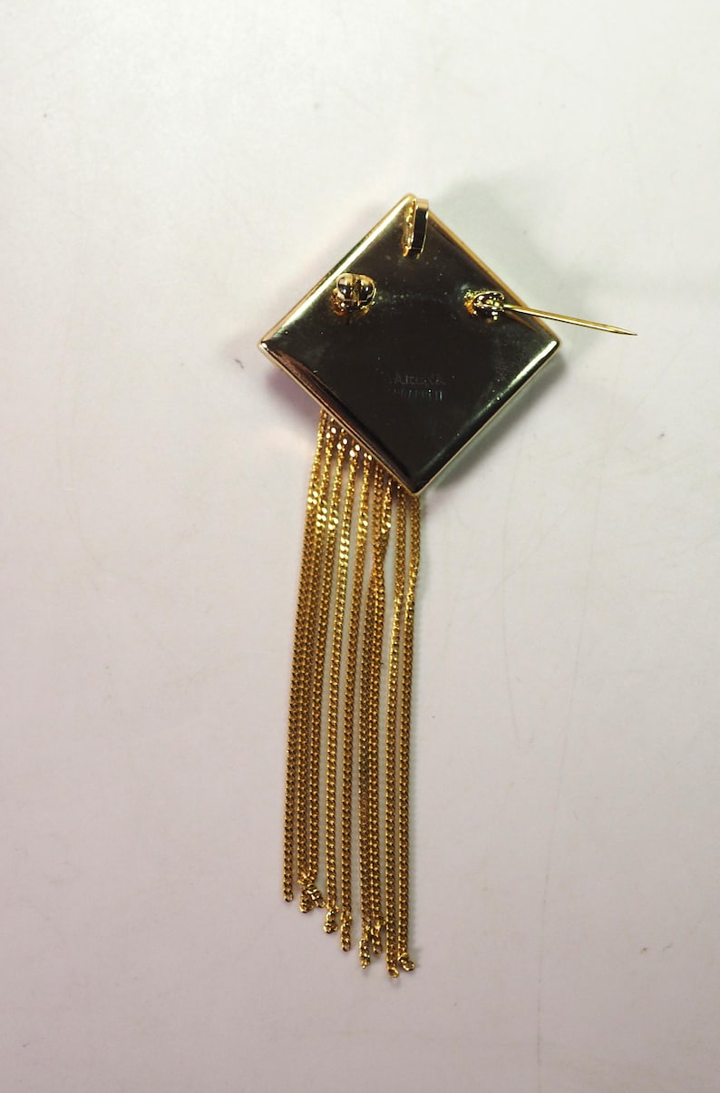 May include: A gold-toned brooch with a diamond-shaped top and dangling chains. The brooch has a pin closure on the back. The chains are made of small, linked segments. The brooch is likely vintage or costume jewelry.