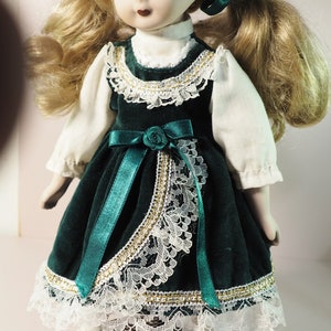 May include: A porcelain doll with blonde hair styled in pigtails, adorned with green ribbons. The doll wears a dark green velvet dress with white lace trim and a matching green bow. The doll is standing on a wooden base.