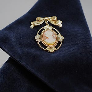 Könnte beinhalten: Eine goldfarbene Brosche mit einem ovalen Kamee, das das Profilporträt einer Person zeigt. Das Kamee ist in einen kreisförmigen Rahmen mit vier floralen Akzenten eingefasst und wird von einer dekorativen Schleife gekrönt, alles in Metalloptik.
