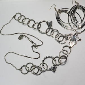 May include: A silver-coloured jewellery set comprising a necklace and earrings. The necklace features a chain with connected rings and butterfly accents. The earrings are large, layered oval shapes. The jewellery is displayed on a white surface.