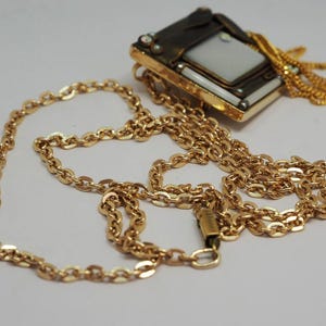 May include: A gold-toned necklace with a rectangular pendant featuring a white center and decorative accents. The chain is a classic link style, and the pendant has a vintage aesthetic. The necklace is displayed on a white surface.