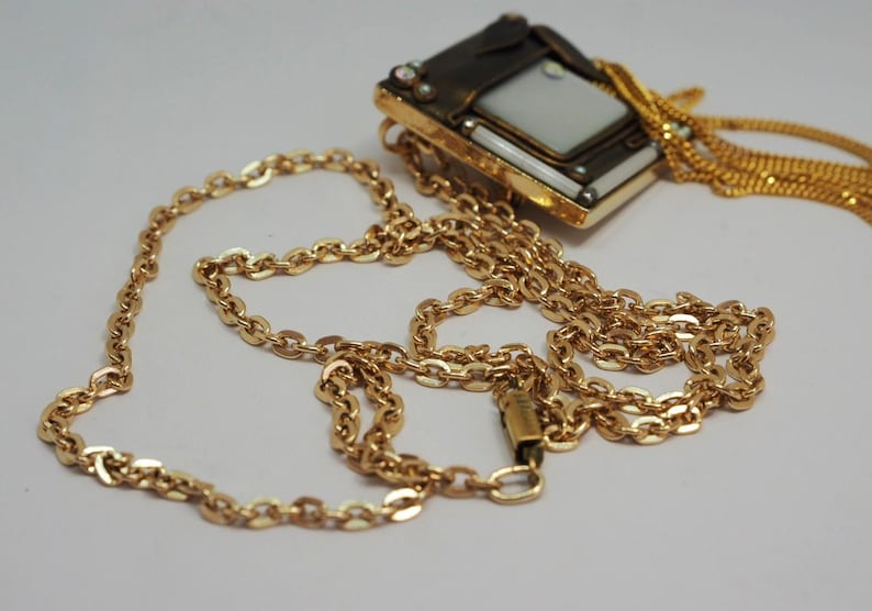 May include: A gold-toned necklace with a rectangular pendant featuring a white center and decorative accents. The chain is a classic link style, and the pendant has a vintage aesthetic. The necklace is displayed on a white surface.
