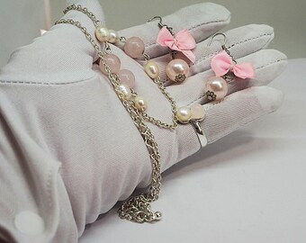 Puede incluir: Un conjunto de collar, pendientes y anillo de plata. El collar presenta una cadena con perlas y cuentas rosas. Los pendientes tienen lazos rosas y grandes gotas de perlas. El anillo tiene un diseño de perlas y cuentas rosas.