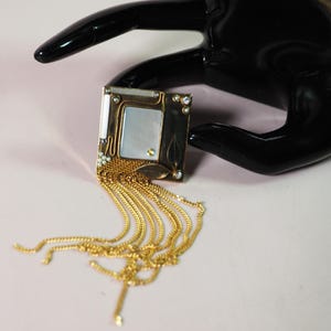 May include: A square, Art Nouveau-style brooch with a gold-tone frame, a mother-of-pearl center, and dangling gold chains. The brooch is embellished with small crystals and leaf accents. The background is a light pink.