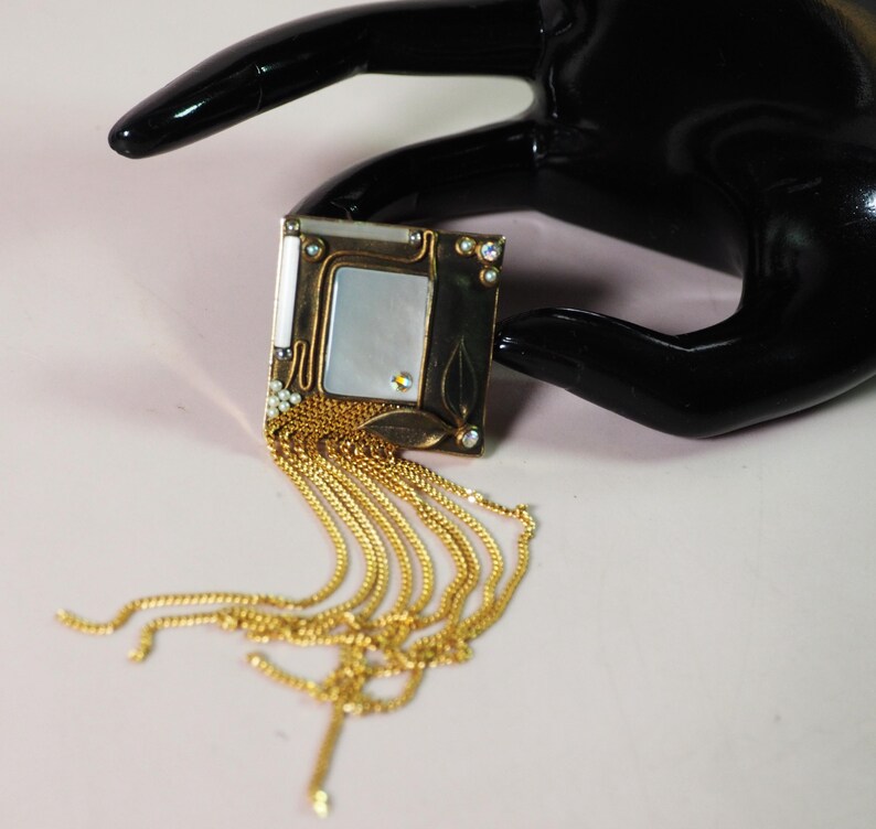 May include: A square, Art Nouveau-style brooch with a gold-tone frame, a mother-of-pearl center, and dangling gold chains. The brooch is embellished with small crystals and leaf accents. The background is a light pink.