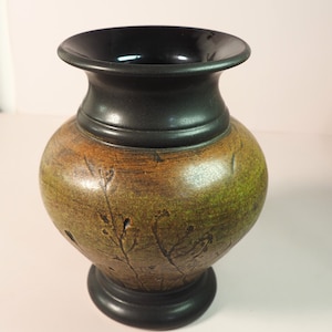 Puede incluir: Un jarrón pequeño de cerámica verde y marrón con un borde y una base negros. El jarrón tiene una superficie texturizada con un diseño de ramas y hojas.