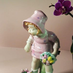 Puede incluir: Una figurita de porcelana de una niña con un vestido y un sombrero rosas, sosteniendo un ramo de flores. La figurita está de pie sobre una base verde con flores.