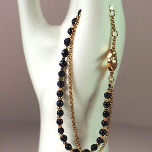 Puede incluir: Una pulsera de cadena dorada con dos hilos. Uno presenta pequeñas cuentas azul oscuro, y el otro es una cadena delicada. La pulsera se abrocha con un cierre dorado.