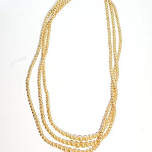 Puede incluir: Un collar de tres hilos con perlas de imitaci&oacute;n de color crema. El collar tiene un cierre plateado y los hilos son de diferentes tama&ntilde;os, con las perlas m&aacute;s grandes en la parte inferior. Se muestra sobre un fondo blanco.