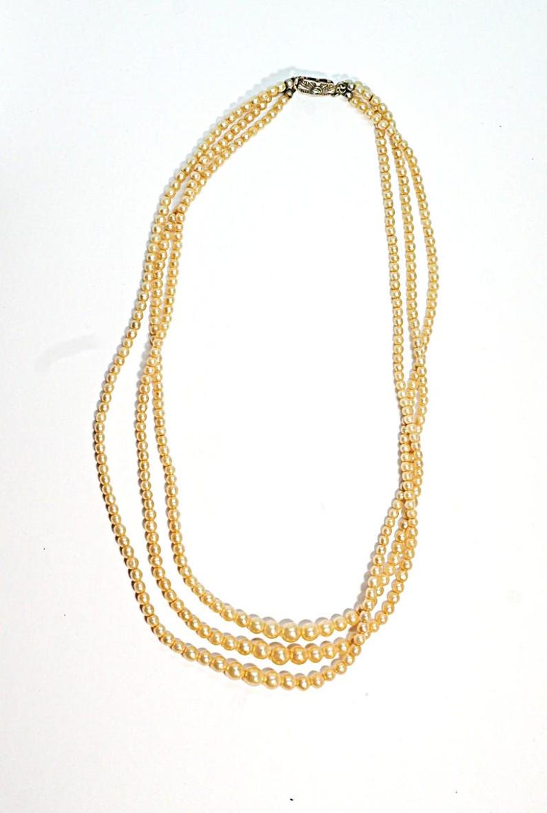 Puede incluir: Un collar de tres hilos con perlas de imitaci&oacute;n de color crema. El collar tiene un cierre plateado y los hilos son de diferentes tama&ntilde;os, con las perlas m&aacute;s grandes en la parte inferior. Se muestra sobre un fondo blanco.