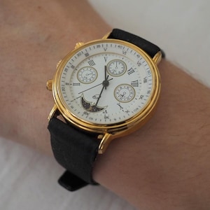 Könnte beinhalten: Eine goldfarbene Armbanduhr mit weißem Zifferblatt, schwarzem Lederarmband und römischen Ziffern. Das Zifferblatt verfügt über drei Hilfszifferblätter, eine Mondphasenanzeige und dünne schwarze Zeiger. Das Gehäuse ist rund und goldfarben.