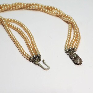 Puede incluir: Un collar vintage con tres hilos de perlas de imitaci&oacute;n color crema. El collar tiene un cierre plateado con un dise&ntilde;o ornamentado y un peque&ntilde;o acento verde. Las perlas est&aacute;n ensartadas, creando un aspecto cl&aacute;sico y elegante.