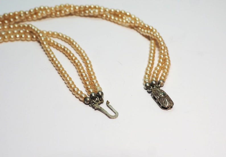Puede incluir: Un collar vintage con tres hilos de perlas de imitaci&oacute;n color crema. El collar tiene un cierre plateado con un dise&ntilde;o ornamentado y un peque&ntilde;o acento verde. Las perlas est&aacute;n ensartadas, creando un aspecto cl&aacute;sico y elegante.