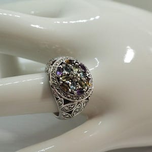 May include: Silver ring with an oval-shaped cluster of colorful gemstones, including purple, green, and clear stones. The ring has a detailed filigree design on the band and around the gemstone setting. The ring is displayed on a white surface.