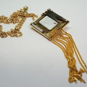May include: Gold-toned necklace featuring a pendant with a square mother-of-pearl center, adorned with small crystals and gold chain fringe. The chain is a delicate, twisted design. The pendant has a geometric shape with a vintage aesthetic.