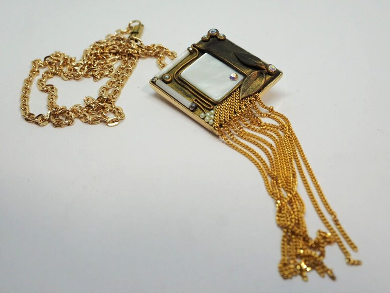 May include: Gold-toned necklace featuring a pendant with a square mother-of-pearl center, adorned with small crystals and gold chain fringe. The chain is a delicate, twisted design. The pendant has a geometric shape with a vintage aesthetic.