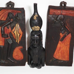 Könnte beinhalten: Drei geschnitzte Holzskulpturen. Zwei rechteckige Wandbehänge zeigen Figuren in Orange und Schwarz. Eine zentrale schwarze Skulptur zeigt eine Frauenbüste mit einer Vase auf dem Kopf. Die Schnitzereien haben eine rustikale, tribale Ästhetik.