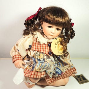 May include: A porcelain doll with brown curly hair styled in pigtails, adorned with red ribbons. The doll wears a dress with a plaid pattern and a decorative apron. A wooden tag reads "Adele Puppen".