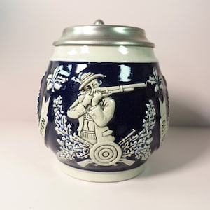 Peut inclure: Une chope à bière en céramique bleue et blanche avec un couvercle en étain. La chope présente un motif en relief d'un homme tenant un fusil, entouré de feuilles et d'une cible. Le texte "Gut Schuss" est visible sur la chope.