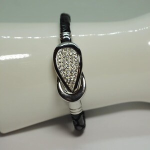 Puede incluir: Una pulsera de cuero negro con un diseño de nudo plateado. El nudo está incrustado con pequeños cristales transparentes. La pulsera se muestra sobre una superficie blanca y curvada, resaltando su elegante diseño y detalles brillantes.