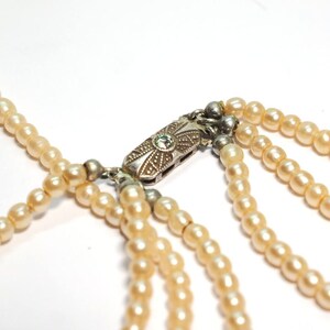 Puede incluir: Primer plano de un collar de perlas de varias hebras. El collar presenta m&uacute;ltiples hebras de perlas de color crema, conectadas por un cierre plateado con un dise&ntilde;o ornamentado y una peque&ntilde;a gema verde. Las perlas son de tama&ntilde;o uniforme y tienen un acabado liso y brillante.