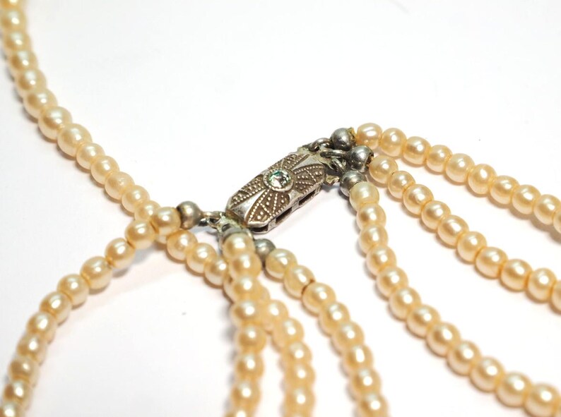 Puede incluir: Primer plano de un collar de perlas de varias hebras. El collar presenta m&uacute;ltiples hebras de perlas de color crema, conectadas por un cierre plateado con un dise&ntilde;o ornamentado y una peque&ntilde;a gema verde. Las perlas son de tama&ntilde;o uniforme y tienen un acabado liso y brillante.